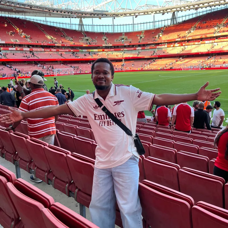 Ibukun at the Emirates Stadium in a white Arsenal shirt, arms spread wide in front of rows of red seats