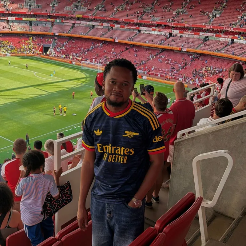 Ibukun in a navy Arsenal shirt in the upper tier of the Emirates Stadium, with the pitch behind