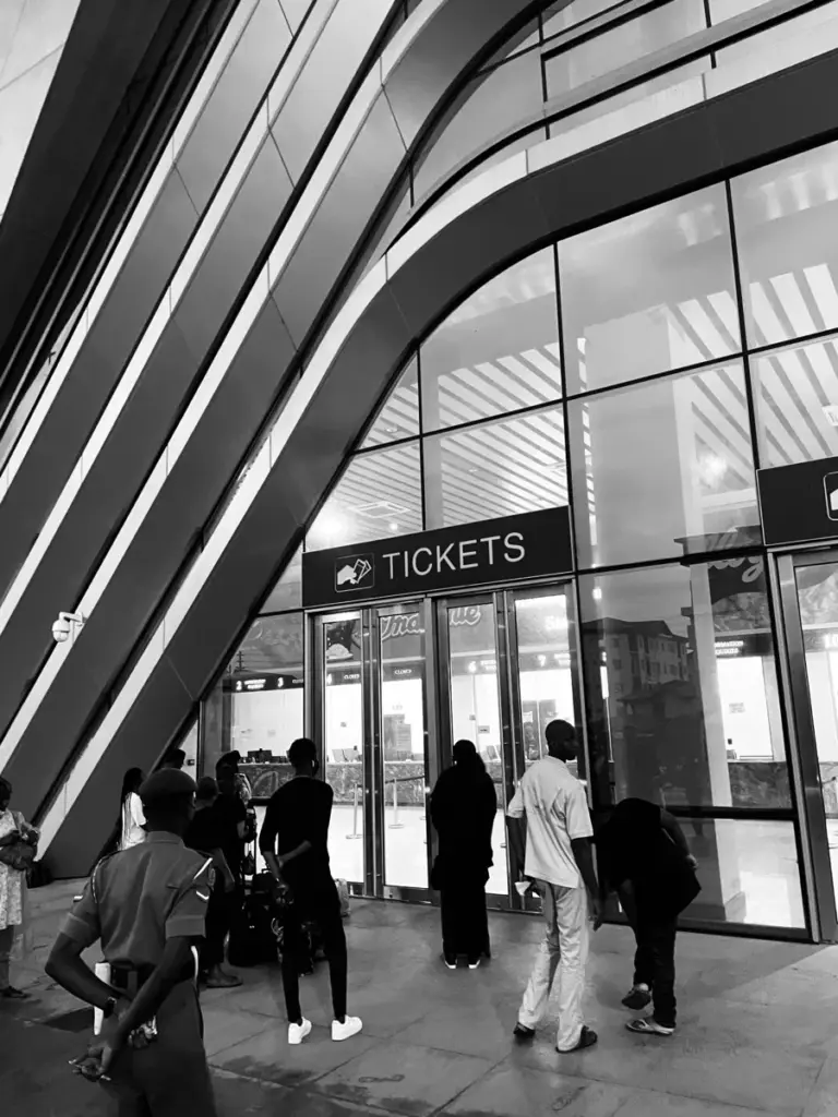 Black and white shot of a curved station front with a Tickets sign and people walking in